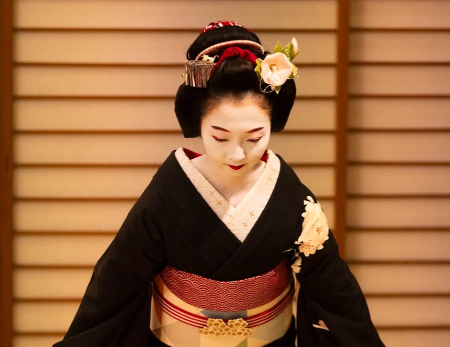 A geisha in a black kimono and red obi kneels before paper screens
