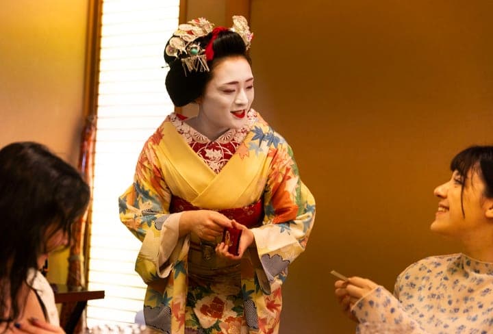 An apprentice geisha greets smiling guests at dinner