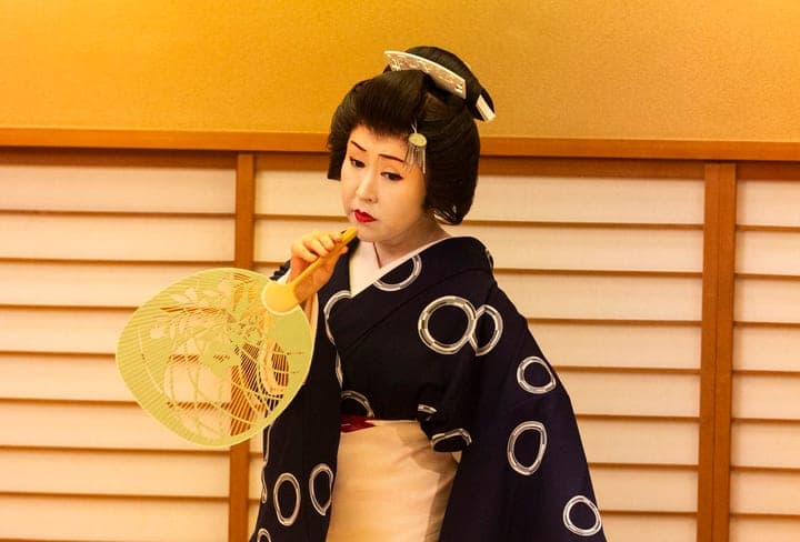 A geisha in a patterned kimono performs with a fan before paper screens