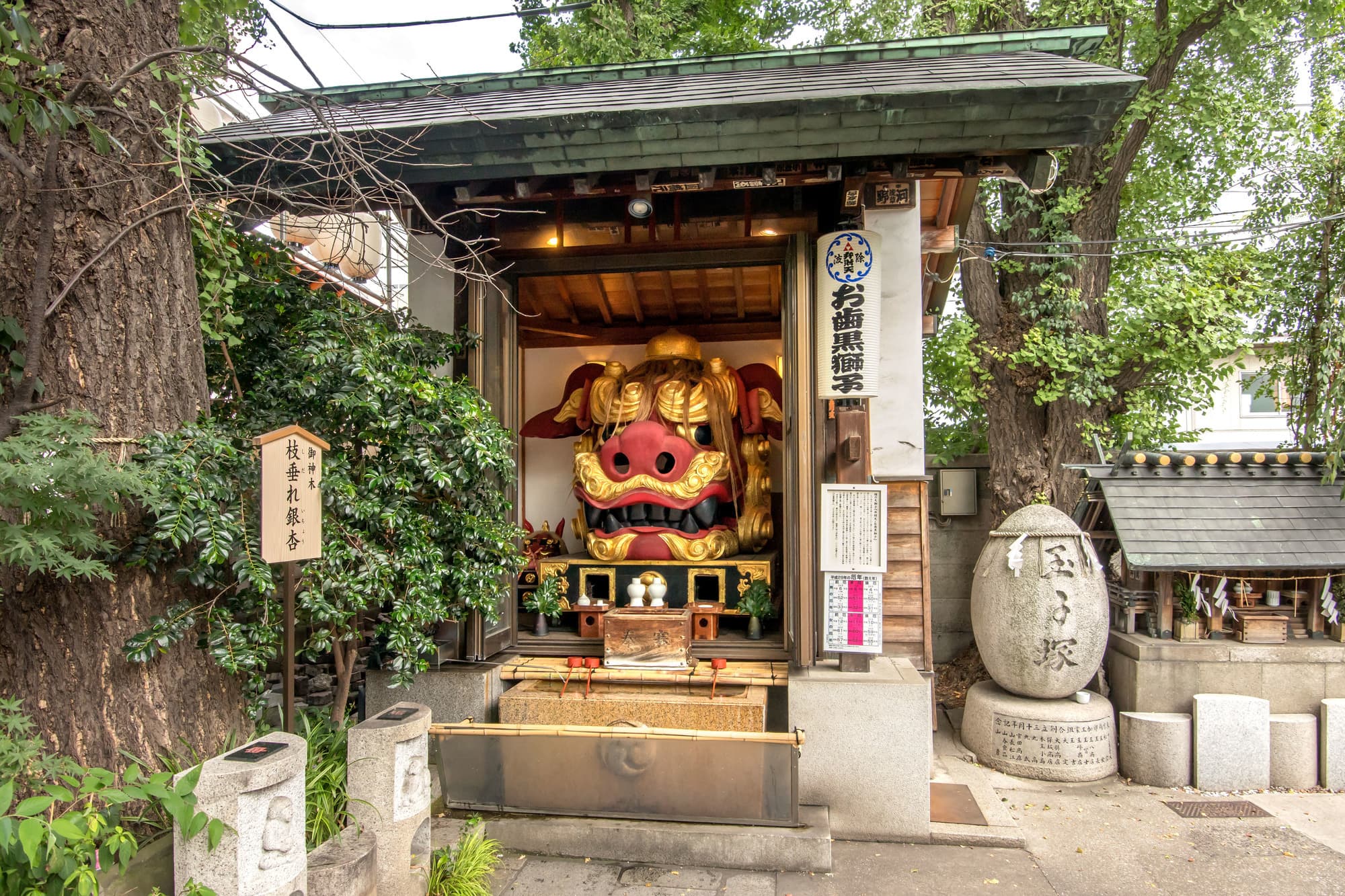 Small Japanese shrine pavilion framed by trees, displaying a vivid red-and-gold lion mask, stone lanterns, signs, and a serene courtyard.