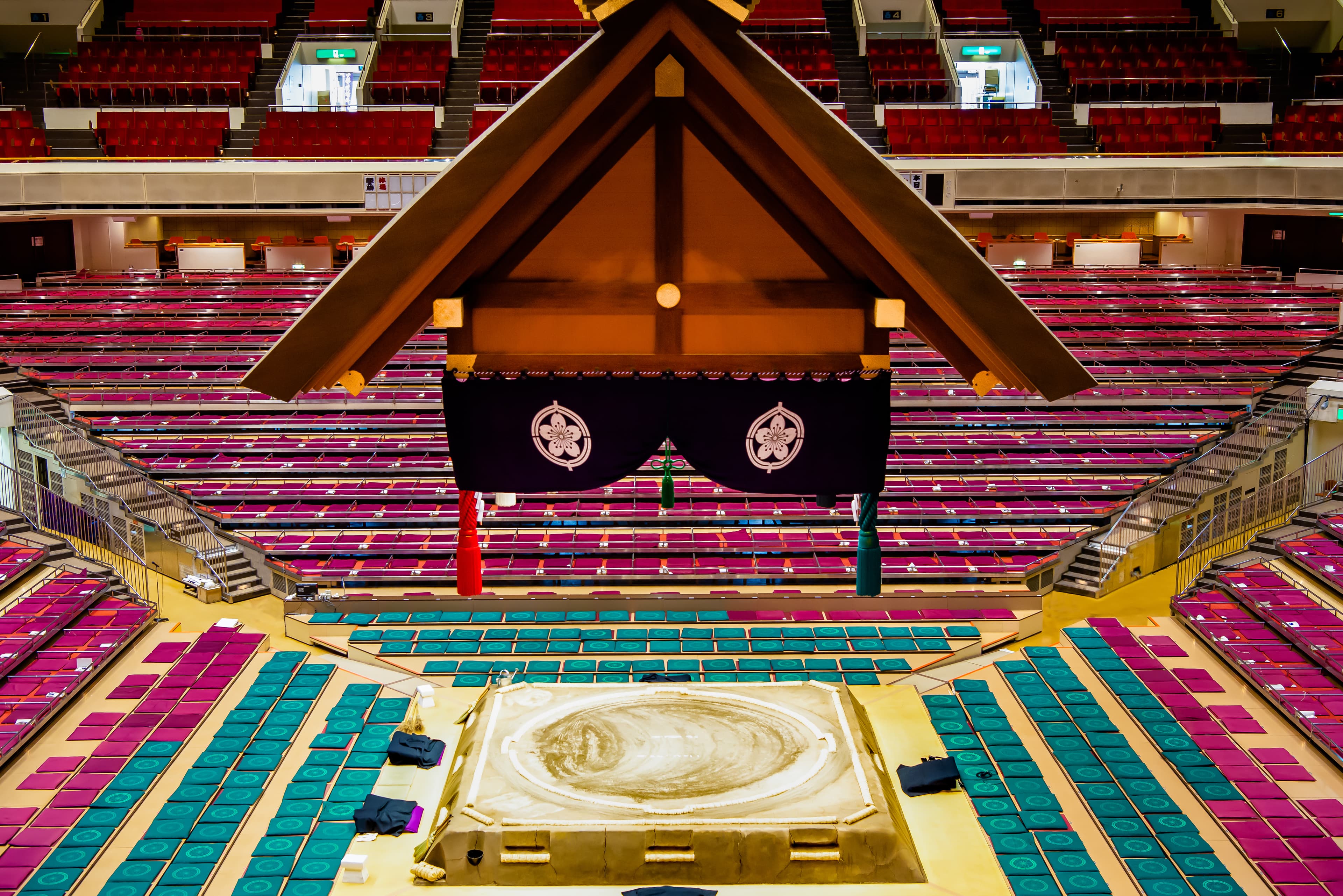 Shrine-style roof over the empty sumo ring at Ryogoku Kokugikan, ringed by colorful cushion seating